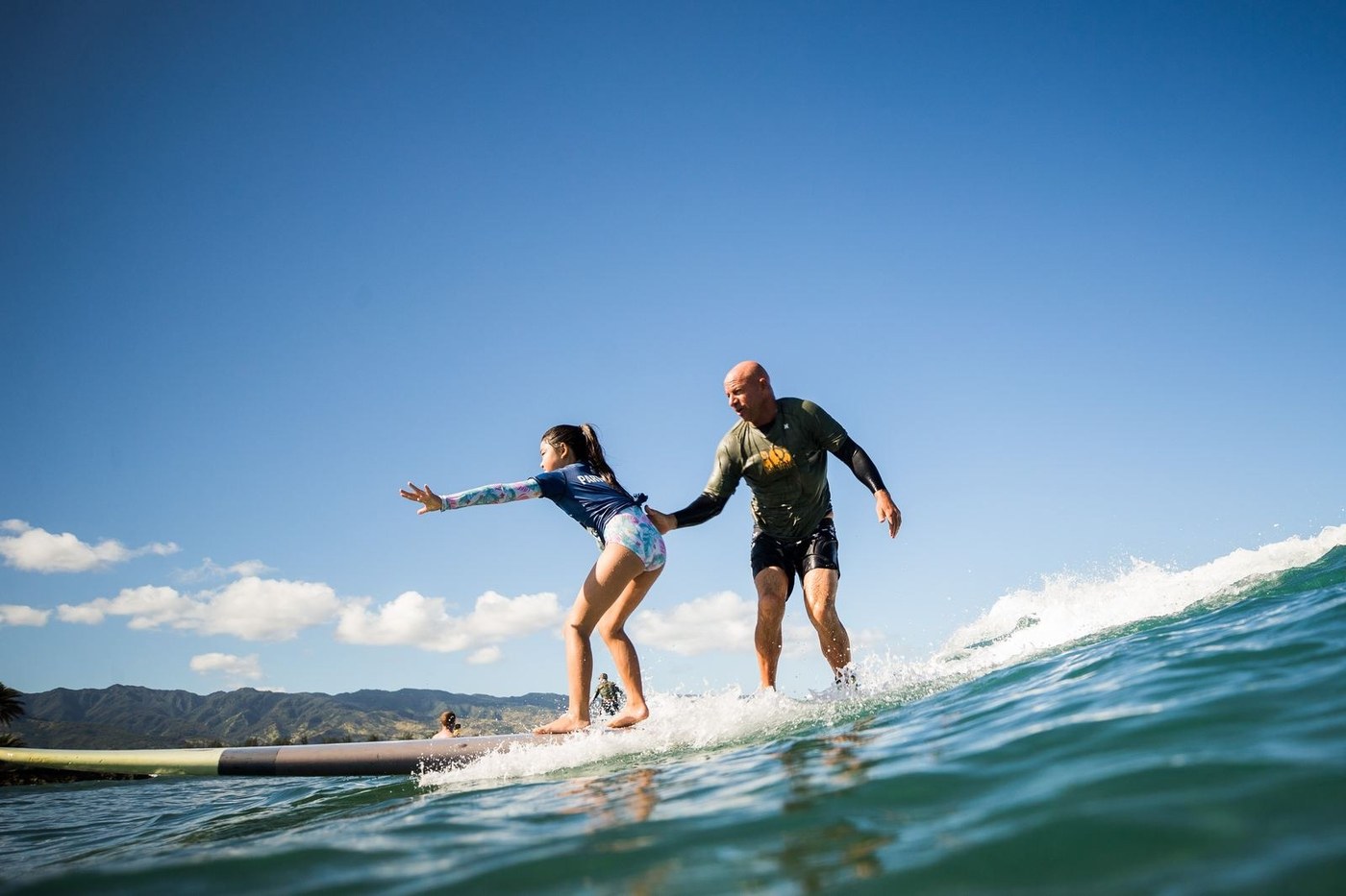 A young participant stands on a wave, arms out, at a Mauli Ola Surf Experience Day