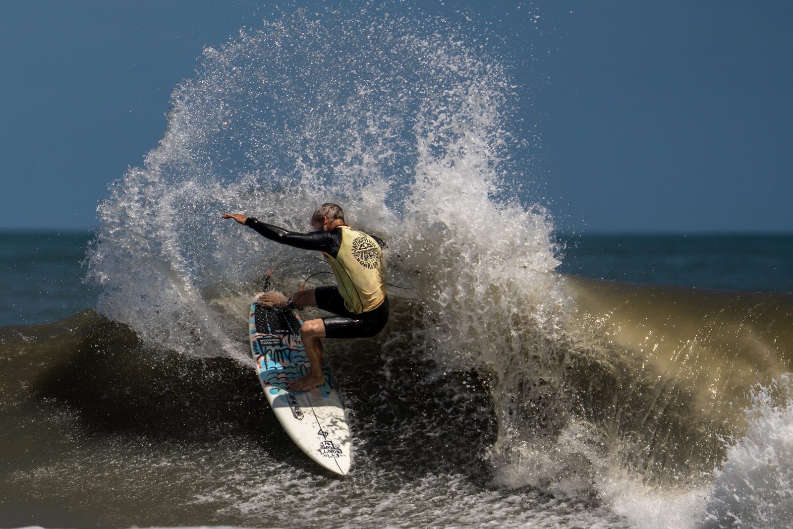 Surfer in a yellow contest jersey throwing a backside hack with a fan of whitewater