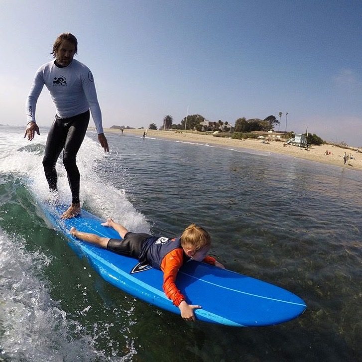 Hans Hagen riding a wave standing behind a young participant lying on the front of a blue soft-top surfboard