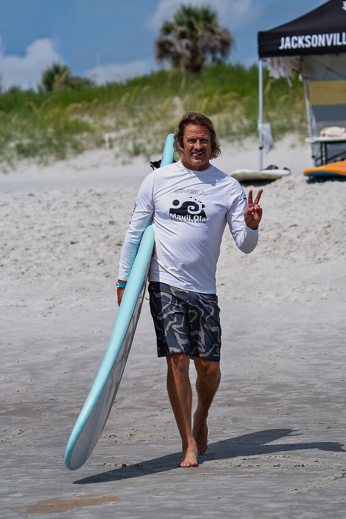 A Mauli Ola Foundation volunteer walks up the beach carrying a soft-top board, flashing a peace sign, a Jacksonville Board Riders tent behind him.