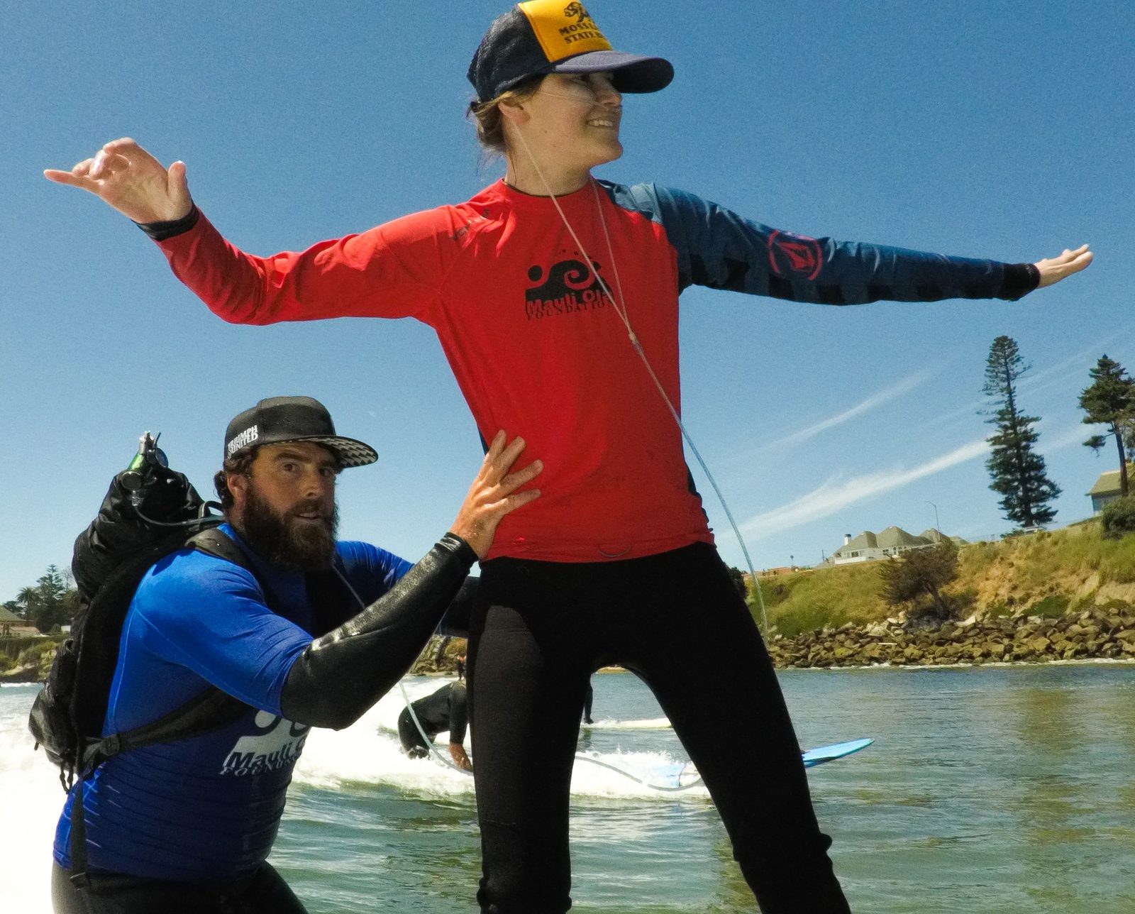 Melissa Pappageorgas standing on a surfboard, arms out, while her husband Joey supports her, carrying her oxygen on his back