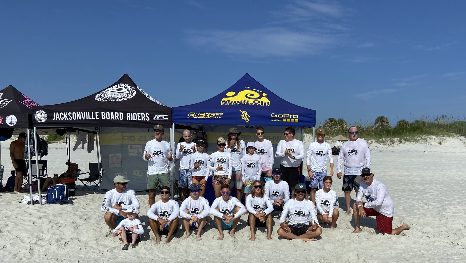 The Mauli Ola Foundation's blue and yellow tent on the sand beside a Jacksonville Board Riders tent, with volunteers in white rash guards gathered in front