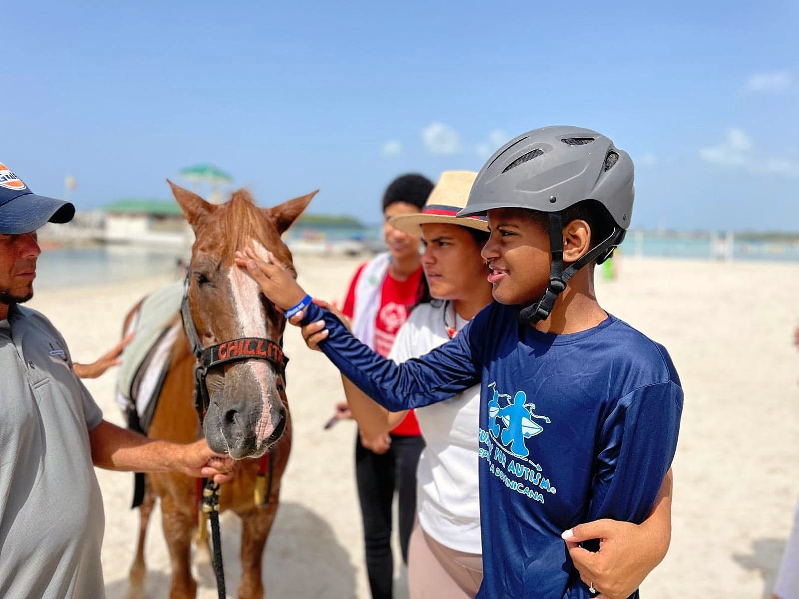 A young athlete in a helmet and a Surfers for Autism República Dominicana jersey reaches out to pet a horse on a bright Dominican beach