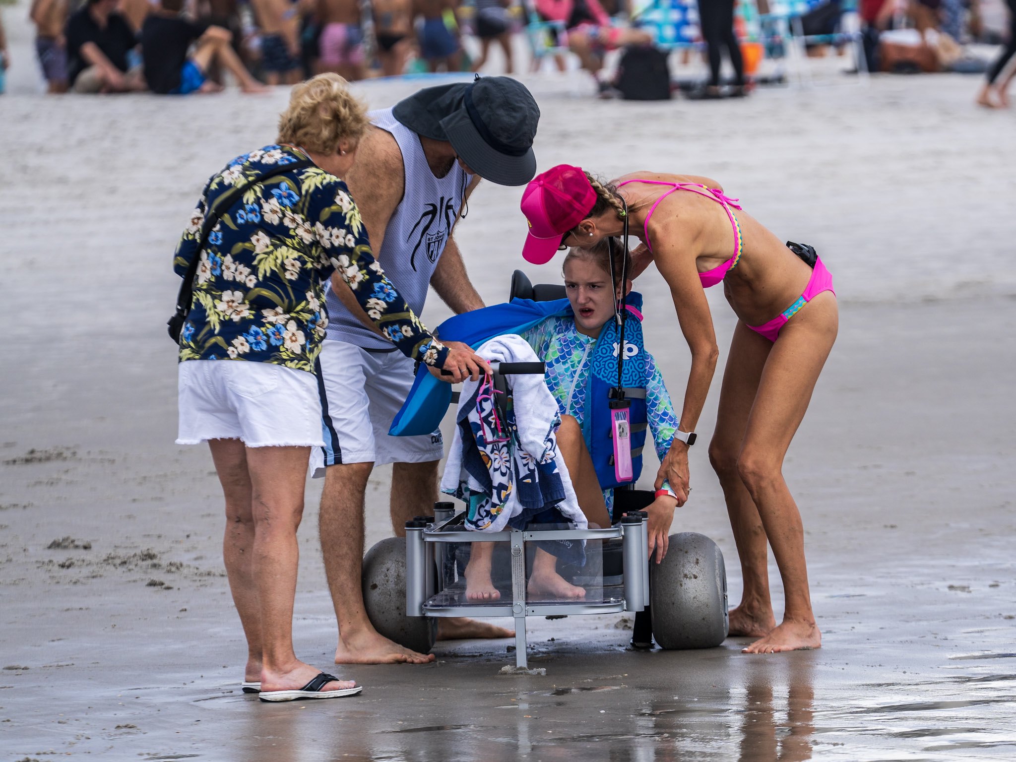 An athlete sits in a surf chair in the shallow water while three Surfers for Autism volunteers gather around to help.
