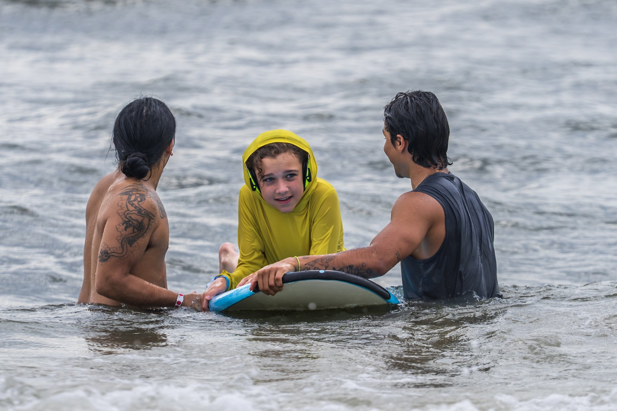 Two volunteers steady a blue surfboard as a young athlete wearing ear defenders looks toward the camera in the water.