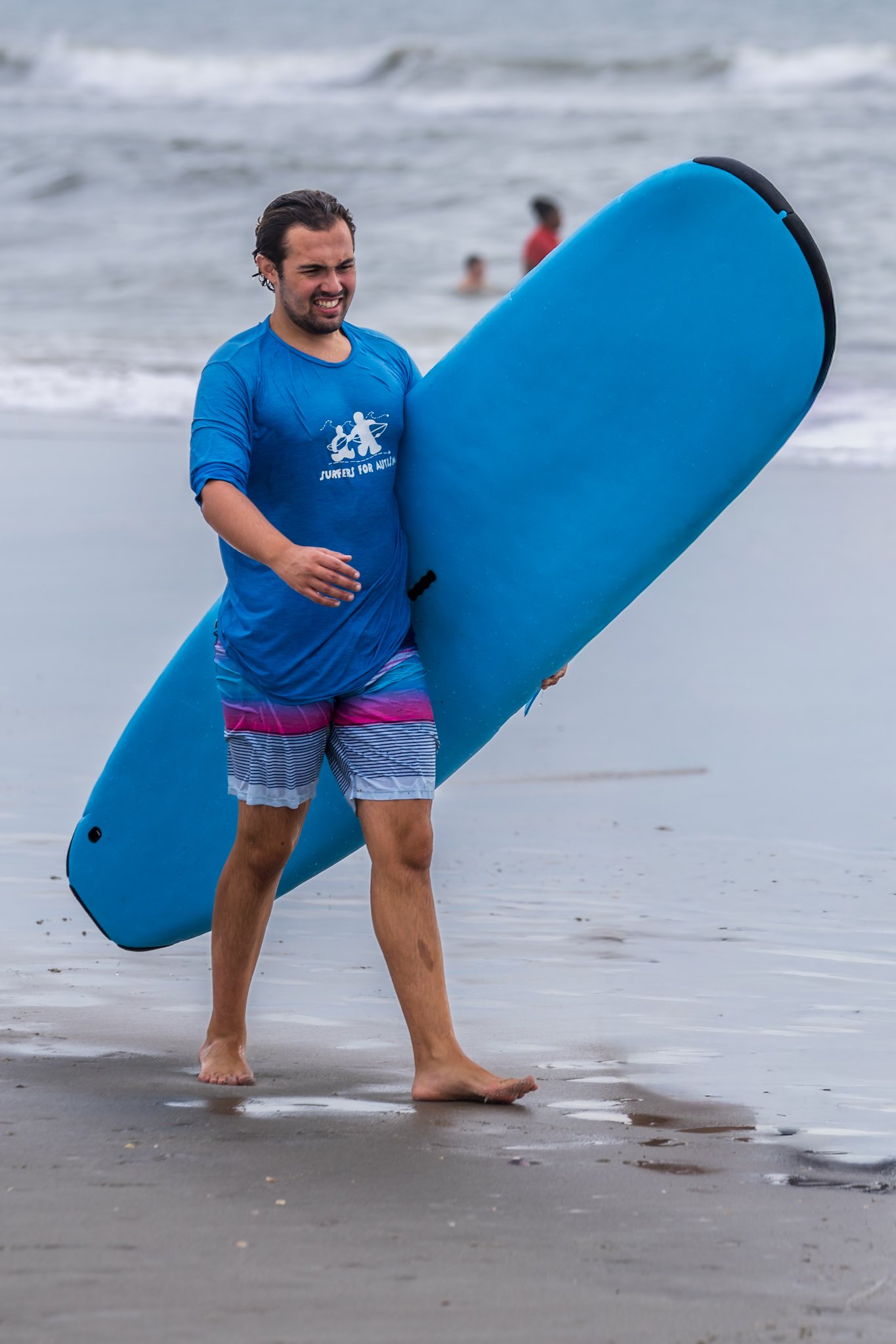 A soaked Surfers for Autism volunteer laughs between sessions while standing beside a surfboard.