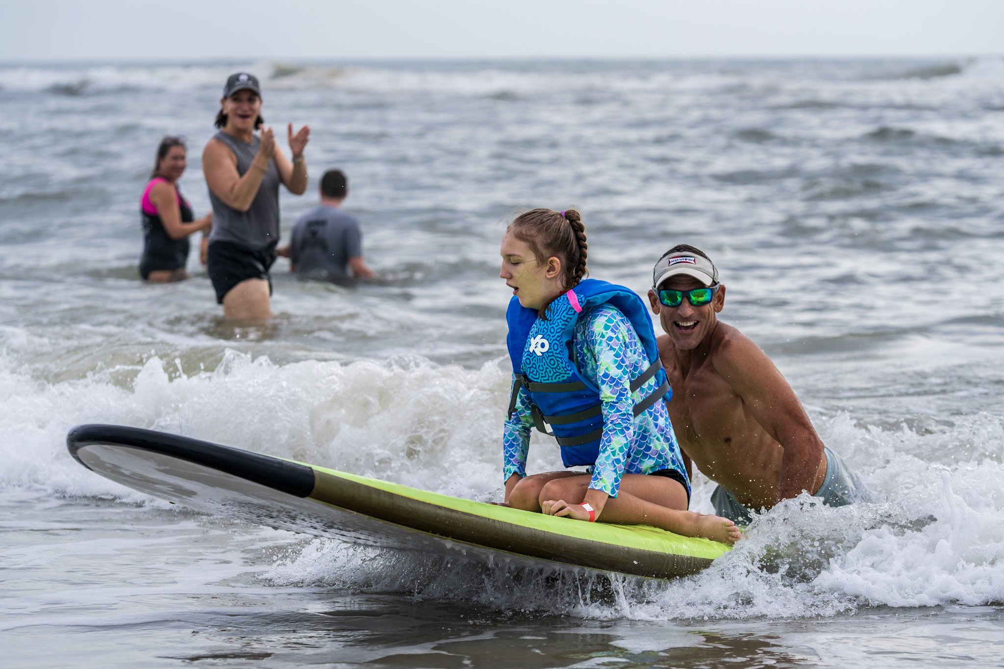 An athlete rides through the inside on a surfboard as a smiling coach follows and a volunteer claps from the shallows.