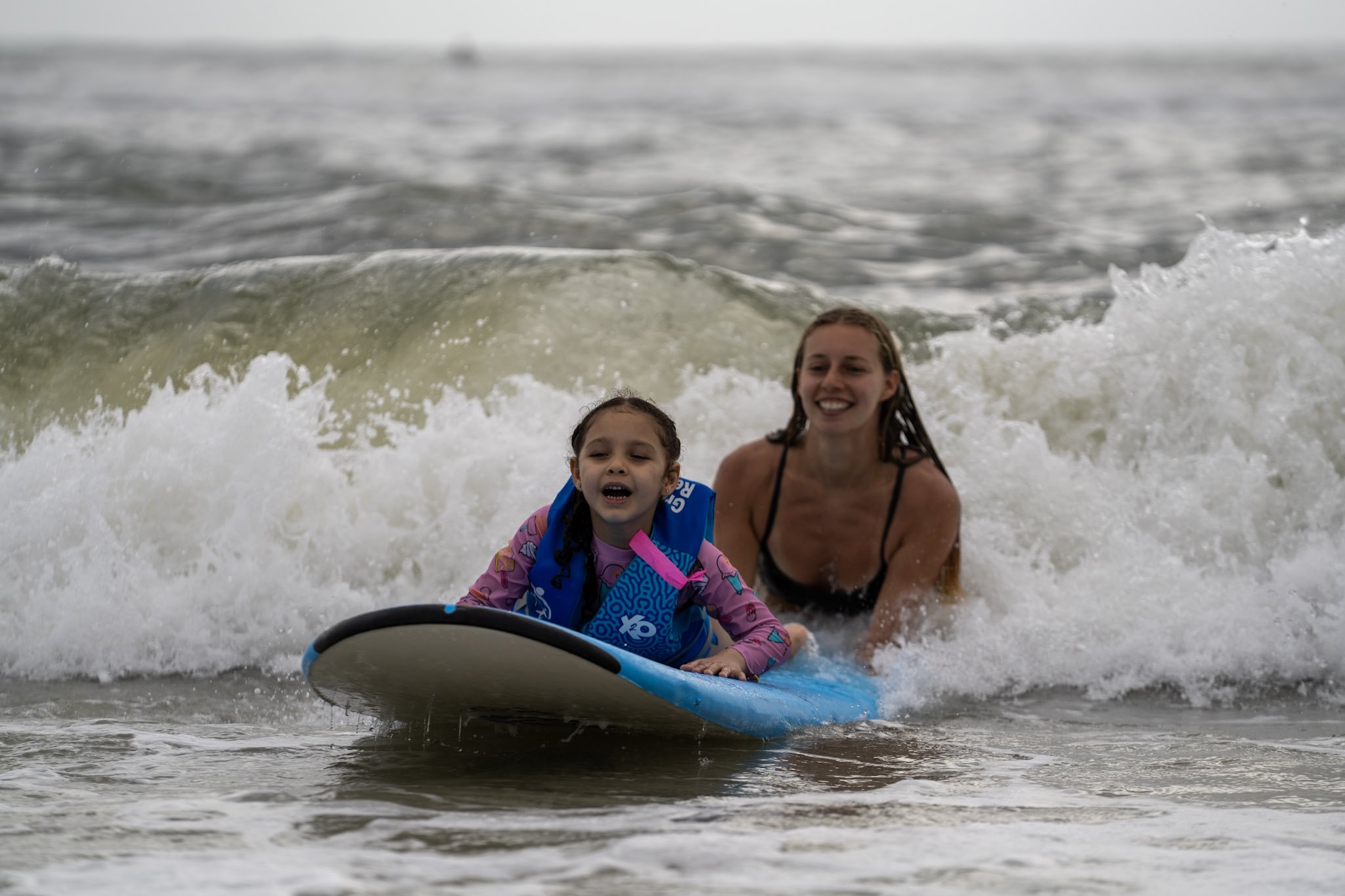 A young athlete and a smiling volunteer ride a blue surfboard through breaking whitewater toward Jacksonville Beach.