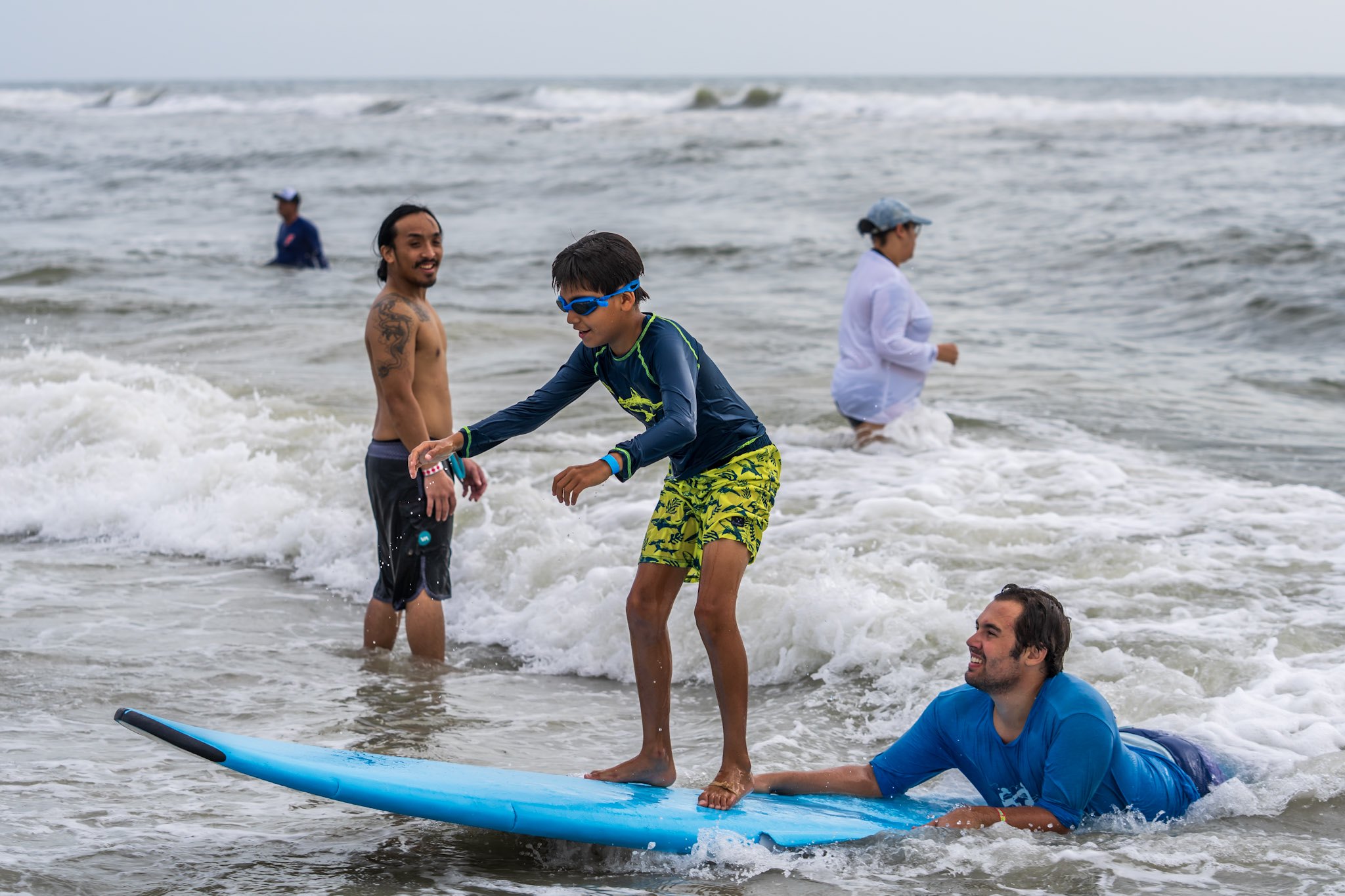 A young athlete wearing goggles stands on a surfboard while Surfers for Autism volunteers remain close in the water.