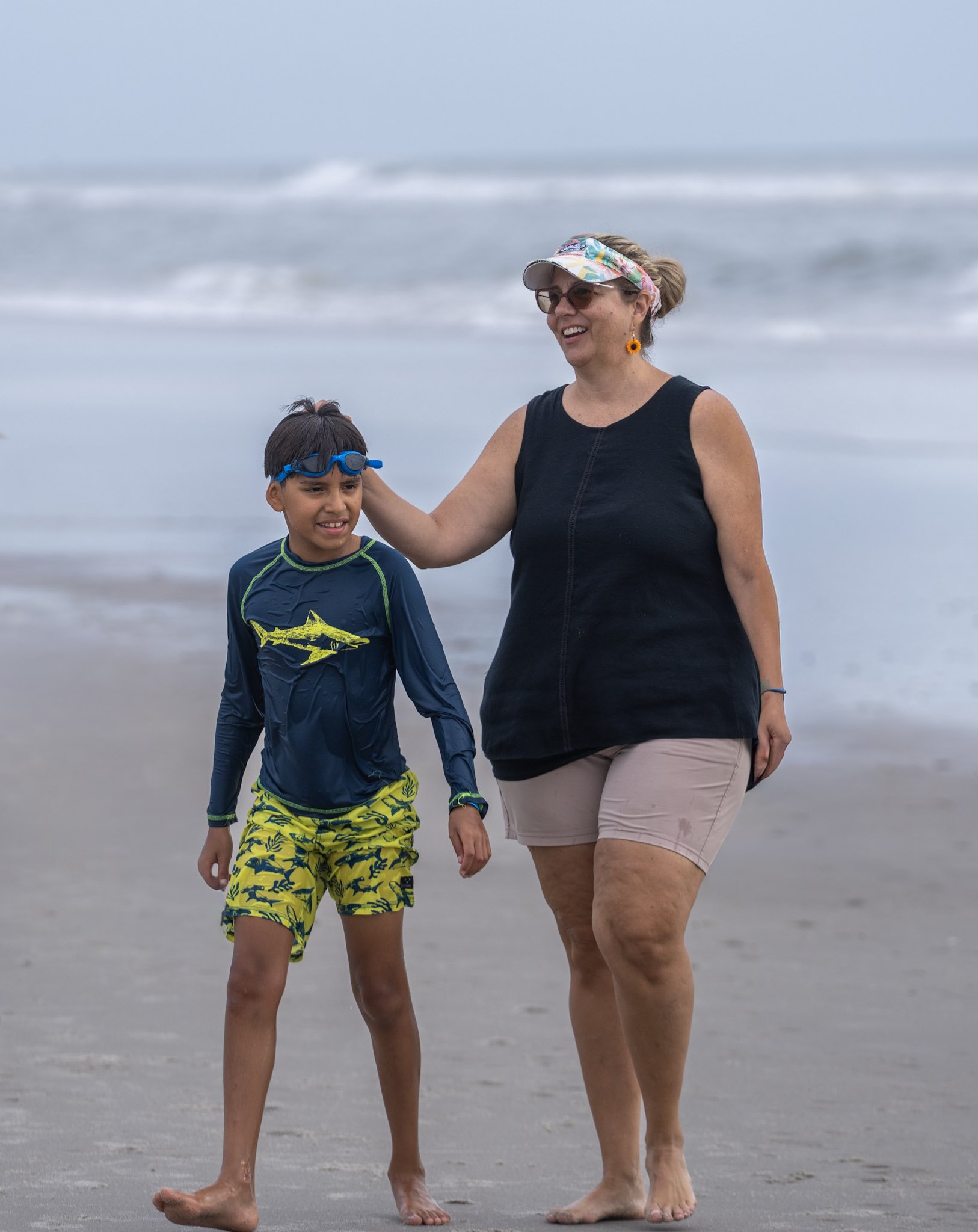 A boy wearing swim goggles stands beside his mother, whose hand rests warmly on his shoulder at the Surfers for Autism event.