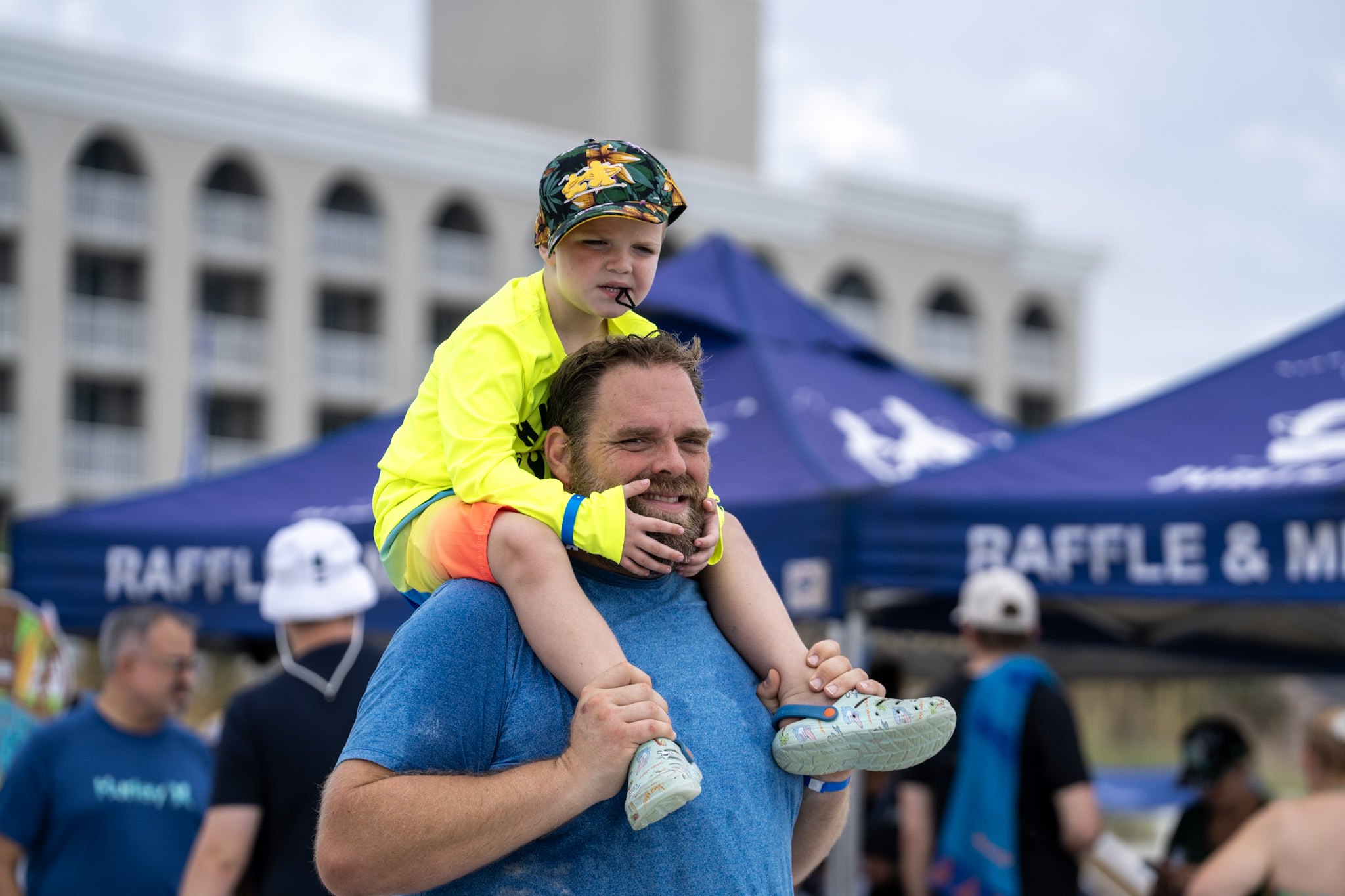 A smiling child sits on a parent's shoulders with Surfers for Autism tents visible behind them on Jacksonville Beach.