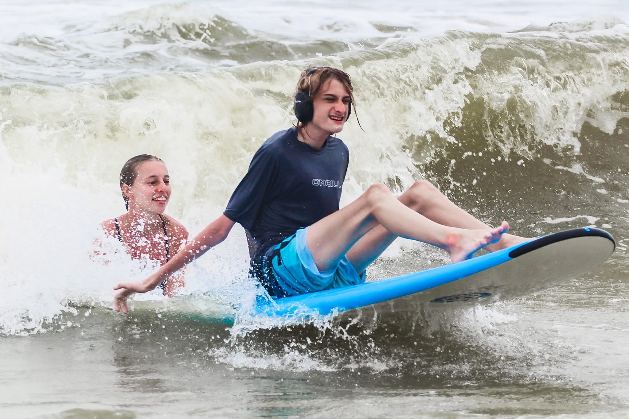 A young athlete in noise-canceling headphones rides the whitewater seated on a blue soft-top surfboard while a smiling volunteer surfaces beside him