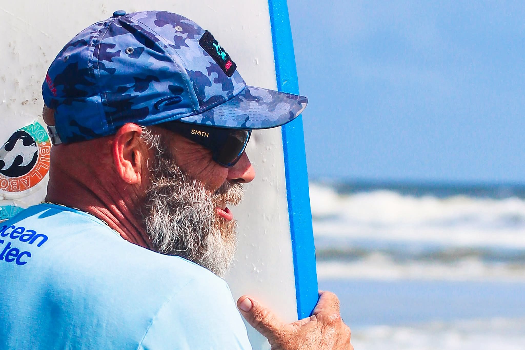 Moose Brown in profile, gray-bearded in a blue camo cap and sunglasses, holding a soft-top surfboard upright and looking out at the surf