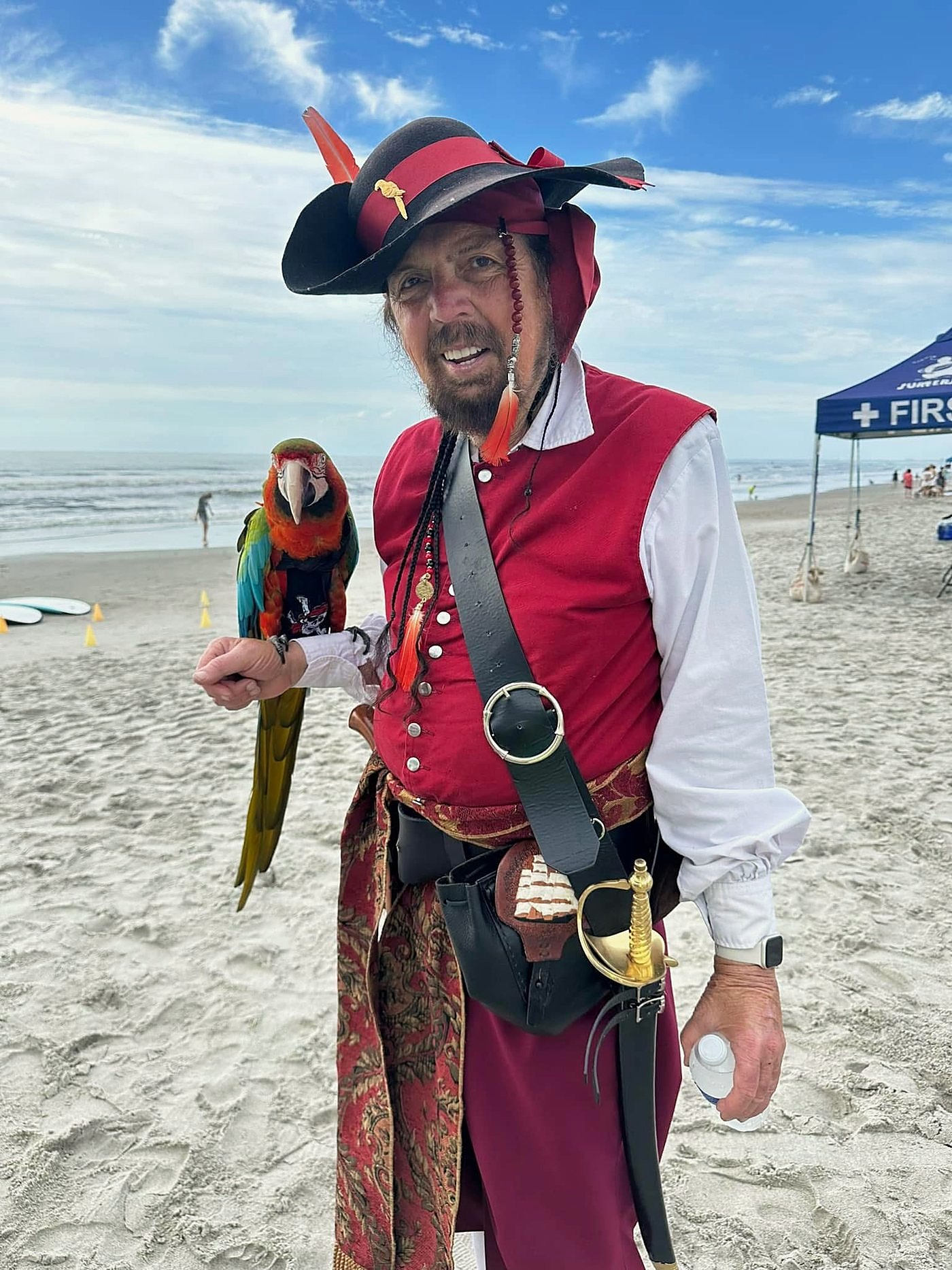 A man in full pirate costume — feathered hat, red vest, cutlass — holds a macaw on his wrist on the beach, a first aid tent and soft-top surfboards behind him