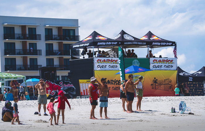 Spectators stand on the sand in front of the Sunrise Surf Shop judging scaffold and sponsor tents.