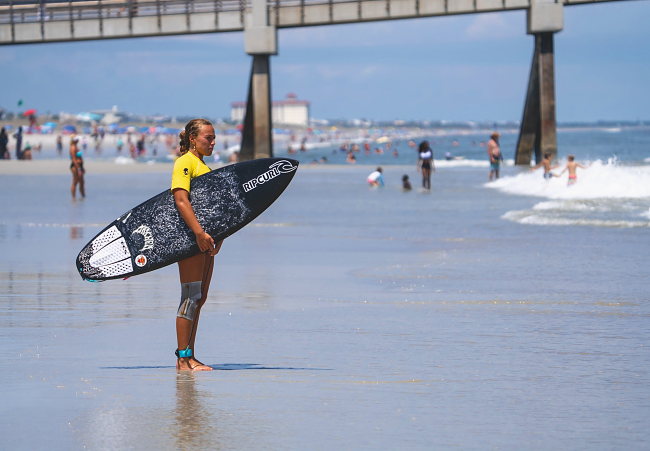 A competitor in a yellow jersey carries a board toward the water.