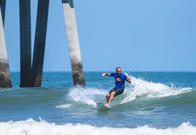 A surfer in a blue jersey rides across a small wave, framed by the pier pilings.