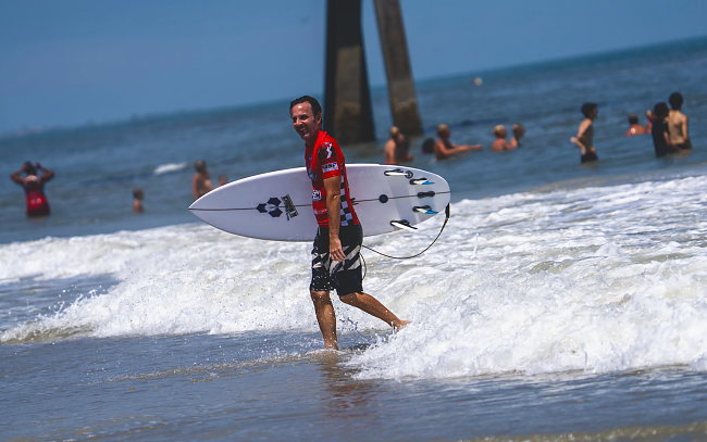 A competitor walks up the beach with a board under one arm, swimmers behind.