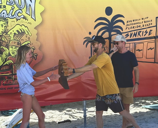 An organizer hands a wooden placing plaque to a young competitor in front of the event banner.