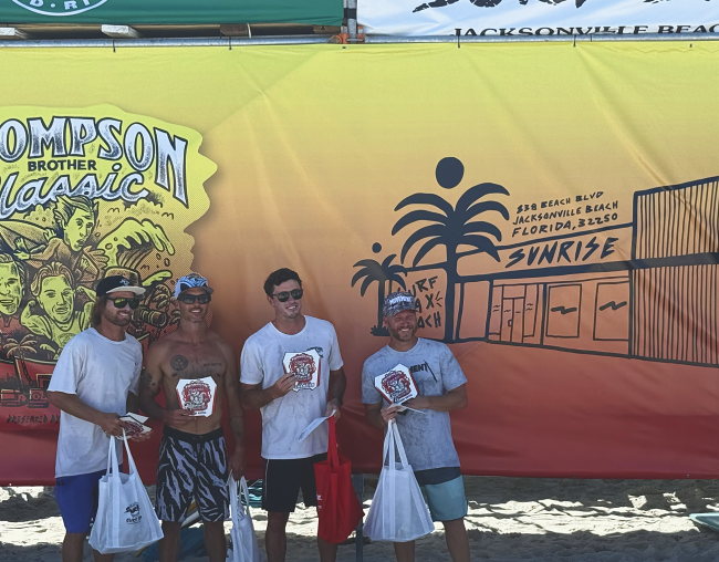 Four men stand in front of the event banner holding placing plaques and prize bags.