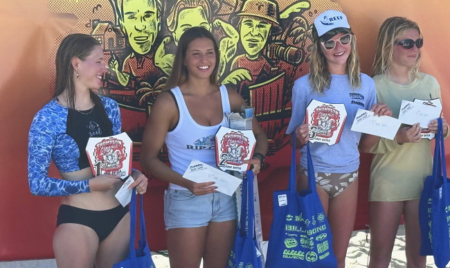 Four women stand in front of the event banner holding placing plaques, envelopes and prize bags.