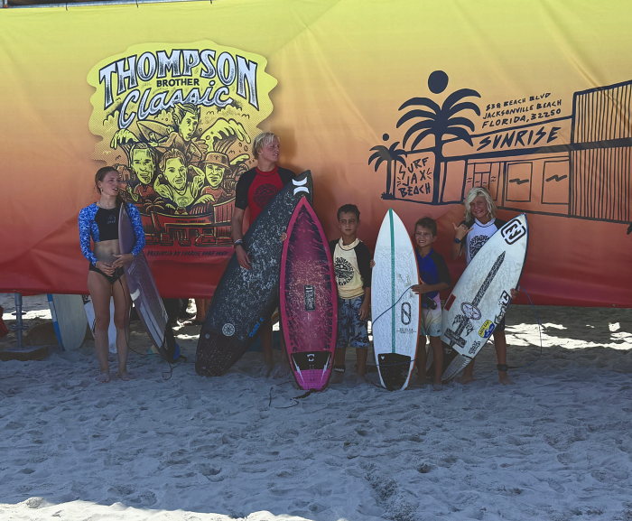 Junior competitors line up with their surfboards in front of the Thompson Brothers Classic banner as results are announced
